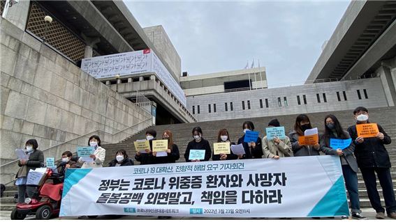 세종문화회관 계단 앞. 코로나19 대확산 전향적 해법 요구 기자회견에 참석한 참가자들이 피켓과 현수막을 들고 있다. 현수막에는 정부는 코로나 위중증 환자와 사망자 돌봄공백 외면말고 책임을 다하라 라고 적혀있다.